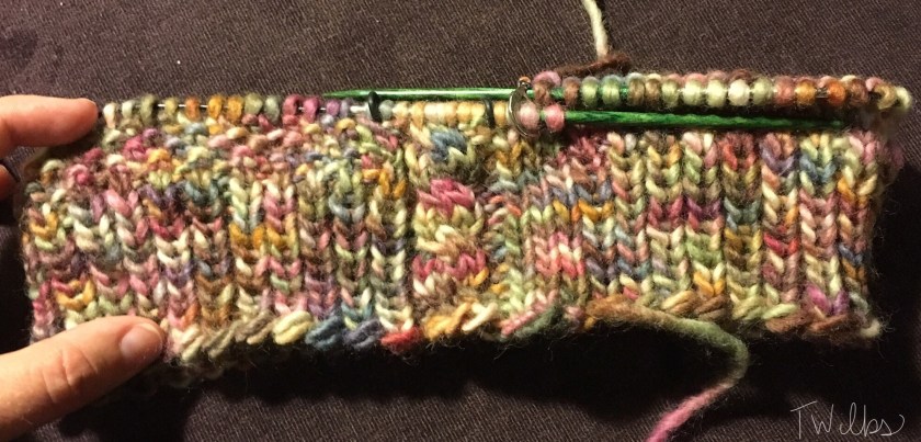 Hand holding open a partially finished hat, still on circular needles, knitted in variegated yarn. There is a cable on the right side, ribbing on the majority of the bottom, and moss stitch to the left. 